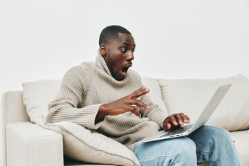 Surprised young African man in a sweater reacting to news on a laptop at home, captured in a light...