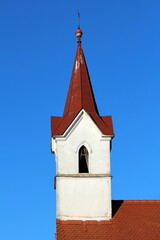 White church steeple with a tall, pointed red spire and weathered paint rises above a tiled roof,...