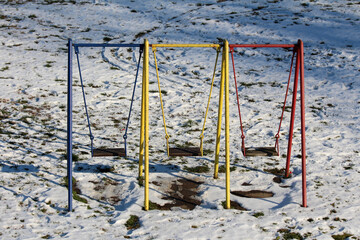Blue, yellow, and red metal swingset with three empty swings stands on a playground covered in patchy snow, with visible footprints and grass beneath the thawing surface