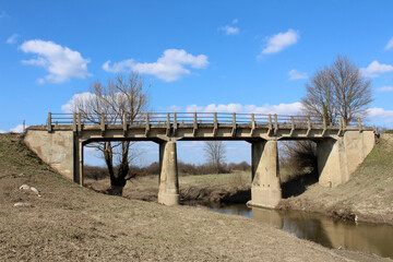 Sturdy concrete bridge with railings spans a narrow river, set in an open grassy landscape with...