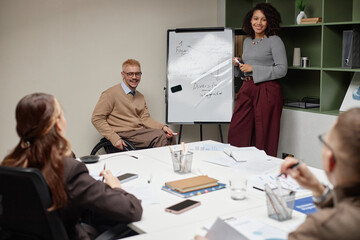 Caucasian man with disability sitting in wheelchair and Black young adult woman standing by whiteboard leading business meeting with diverse colleagues listening
