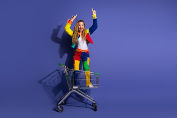 Stylish young woman in colorful casual outfit posing playfully in a shopping cart on a vibrant...