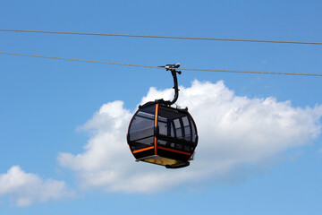 Contemporary gondola lift cabin with tinted windows and orange accents hangs from a steel cable, silhouetted against a bright blue sky and white clouds, used for mountain or urban transport