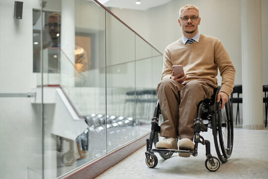 Caucasian young adult man with disability sitting in wheelchair holding smartphone in modern building, looking forward with neutral expression, short blond hair and glasses visible
