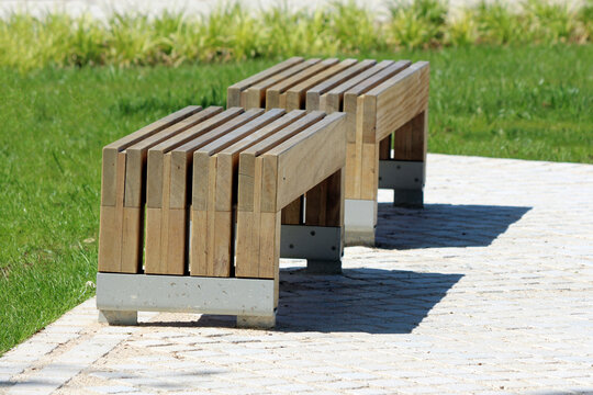 Two minimalist wooden benches with parallel slats and metal supports are positioned on a sunlit stone-paved path, surrounded by green grass and ornamental plants in a public park setting