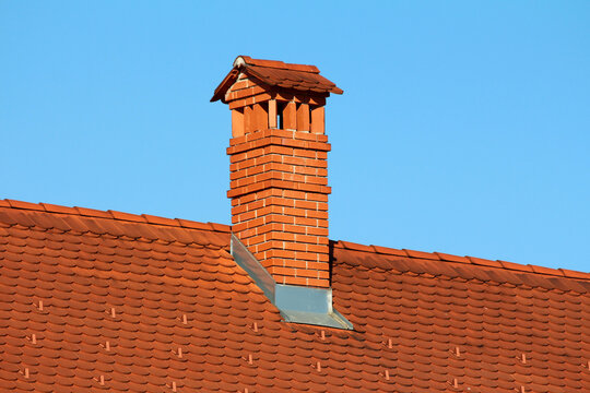 Tall red brick chimney with decorative cap rises from a red clay tile roof, with crisp flashing and regular tile pattern, set against a cloudless blue sky in bright sunlight
