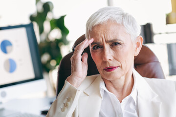 Confident senior businesswoman in formal attire seated in modern office setting with visuals of...