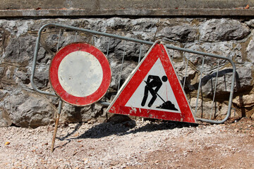 Two weathered road signs—a faded no entry circle and a triangular roadwork warning—lean against...