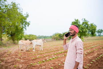 young indian farmer standing at agricultural field talking on smartphone