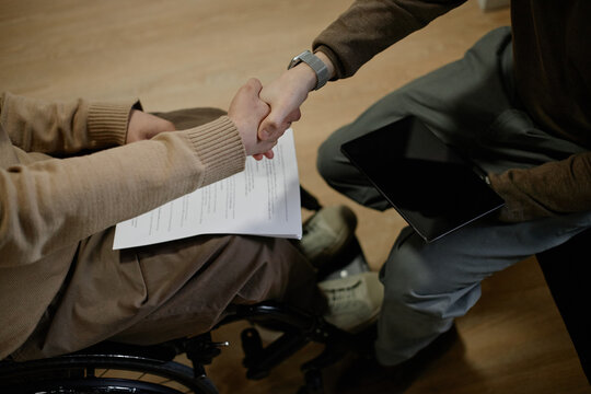 Man with disability sitting in wheelchair shaking hands with partner holding digital tablet during business meeting indoors - Powered by Adobe