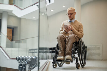 Caucasian young adult man with disability sitting in wheelchair using smartphone in modern building, looking at screen with focused expression