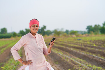 young indian farmer holding smartphone standing in agriculture field