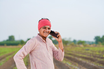 young indian farmer standing at agricultural field talking on smartphone