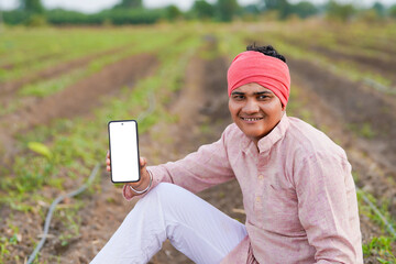 young indian farmer showing smartphone screen sitting at field