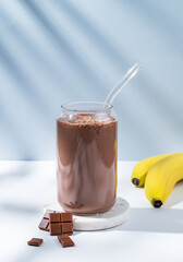 Chocolate smoothie with banana in a glass on a white blue background with morning shadow.