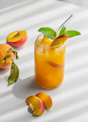 Peach drink or tea with ice, mint in a glass on a white table with fresh fruits and sunlight.