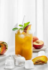 Peach lemonade or tea with mint in a glass on a white table near window with fresh fruits