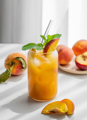 Peach drink or tea with ice, mint in a glass on a white table near window with fresh fruits and sunlight.