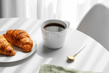 Cup of espresso coffee and fresh croissants on a plate on white table near window with morning...