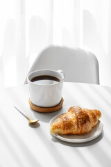 Cup of espresso coffee and fresh croissant on white table near window with morning shadows