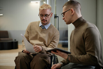 Caucasian young adult man with disability sitting in wheelchair reviewing documents with man...