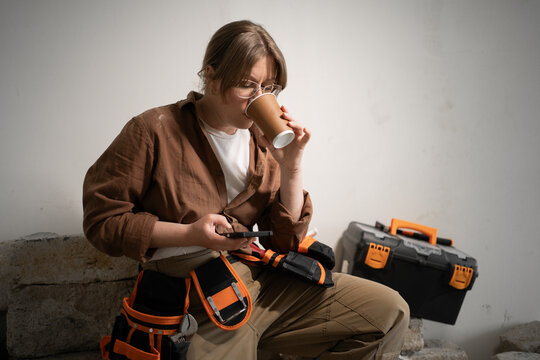 Professional repairwoman in tool belt using smart phone and drinking coffee while resting after work in home