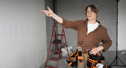 Repairwoman with tool belt in living room interior doing a home renovation, Pointing with finger to space for advertising on gray wall. Renovation and construction concept. Banner.