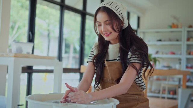 Young asian woman sculpting clay on spinning pottery wheel in ceramic workshop – relaxing creative hobby, art class activity, and handmade design practice in craft studio environment