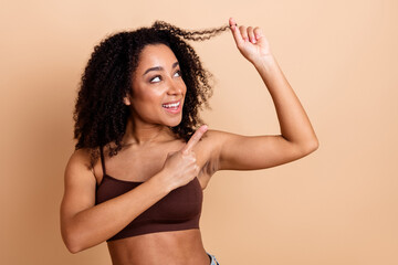 Young woman with curly hair smiles joyfully, pointing to her strand, against a soft beige background