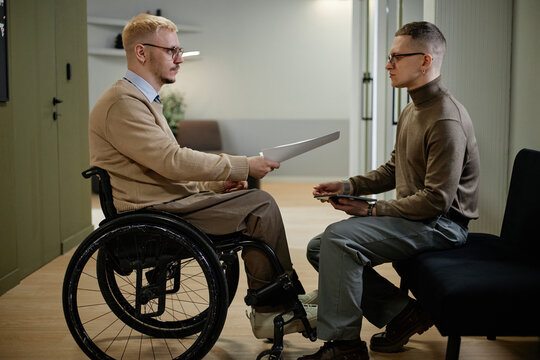 Caucasian young adult man with disability in wheelchair handing document to man sitting opposite, both engaged in business discussion in modern office setting