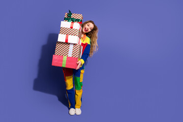 Cheerful young woman in colorful outfit carrying a stack of wrapped gifts against a violet background