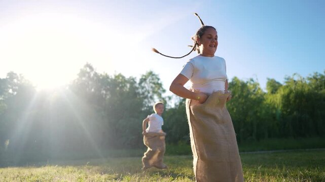 Two women in sack. Children race a sack of grass outdoors. A young girl jumping in the sunlight during a competition in summer. Two women inside a lifestyle bag.