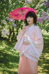 Dreamy portrait of a mysterious geisha-cosplay inspired beauty. Porcelain-skinned woman in a pink ornate Chinese silk robe (Hanfu), short black hair, posing gracefully with Asian parasol on the bloomi