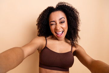 Happy young woman with curly hair in casual brown top taking a cheerful selfie against a beige...
