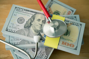 Stethoscope on stack US dollar banknotes with wooden table background. Savings for cost of medical treatment, healthcare, life insurance or personal financial checkup, money saving planning concept.