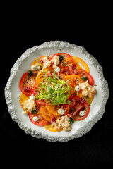 resh summer salad with sliced red and yellow tomatoes, diced vegetables, creamy cheese, herbs, and seeds on a white ceramic plate over a black background. Healthy vegetarian dish.