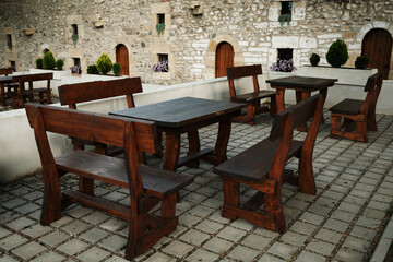 Rustic wooden benches and tables arranged in a stone-paved courtyard against a medieval stone wall with arched doors and flowers. Serbia country.