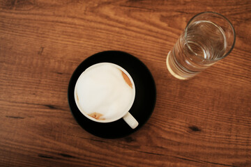 Overhead view of a cappuccino with creamy foam in a white cup on a black saucer and a glass of water on a wooden table. Good morning details at outdoor cafe.