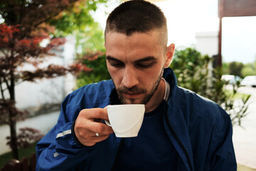 Young man in blue jacket enjoying a cup of coffee outdoors, surrounded by greenery and soft...