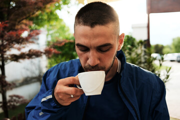 Young man in blue jacket enjoying a cup of coffee outdoors, surrounded by greenery and soft...