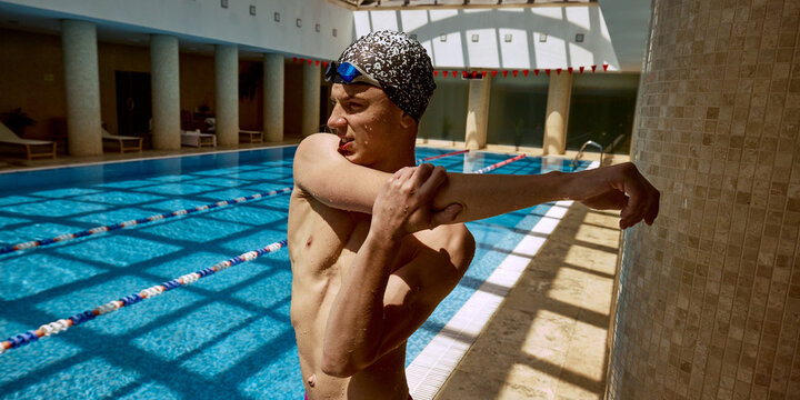 A young swimmer stretches their arm beside an indoor pool under natural light. Concept of pre-swim routines and flexibility for athletes.