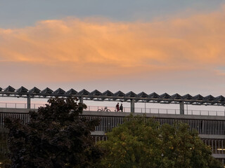 Silhouetted pedestrians with bicycle traverse an elevated modern walkway beneath a dramatic orange sunset sky framed by angular metal canopy structures above leafy trees