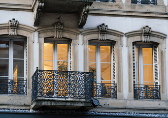 A decorative wrought iron balcony fronts tall wide arched windows glowing warmly from interior lights behind ornate stone facades on a Parisian street