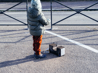 Young child in winter outfit stands beside an empty fireworks box in a quiet parking lot, observing remnants after a New Year festivity