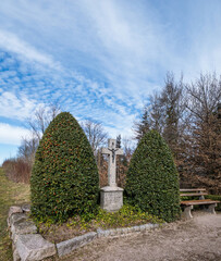 Stone cross monument stands between two trimmed evergreen bushes next to a wooden bench along a rural path in the Black Forest, Germany, under a bright blue sky with scattered clouds