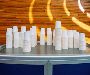 Neatly stacked white paper cups arranged on a metal counter in a modern conference room with warm wooden wall panels and artistic lighting.