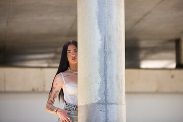 A young, attractive Latin woman with tattoos on her arms and dressed in casual clothes hides behind a concrete column. The woman is wearing make-up.