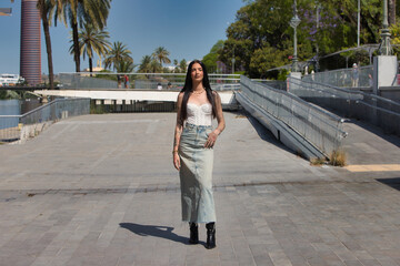 A young, attractive Latin woman with tattoos on her arms and dressed in casual clothes walks and poses on a large city avenue on a sunny day. The woman is wearing make-up.