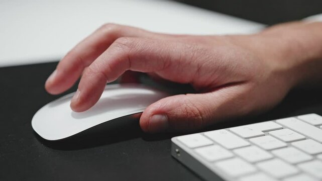 A close-up shot of a person's hand using a modern, white wireless mouse on a black mouse pad. The fingers are actively clicking and scrolling, with a sleek keyboard nearby. Tech and office concepts.