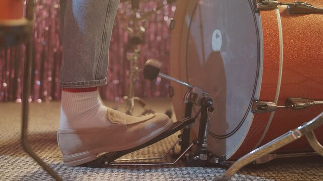 A close-up, low-angle shot of a drummer's foot in a stylish loafer, rhythmically playing the bass drum pedal. The vintage drum and sparkly background suggest a live music performance or band practice.